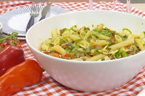 En stor skål med vegansk pastasallad med penne, zucchini, tomater och oliver. Serveras på en rödvitrutig duk.
