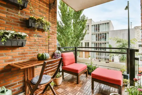 Un balcón alargado decorado con muebles de madera, cojines rojos y plantas colgantes. Un espacio acogedor para disfrutar del exterior.