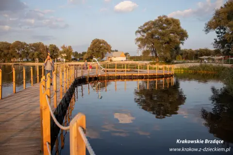 Drewniany pomost nad wodą w największym parku w Krakowie. Ojciec z dzieckiem na ramionach spaceruje po pomoście.