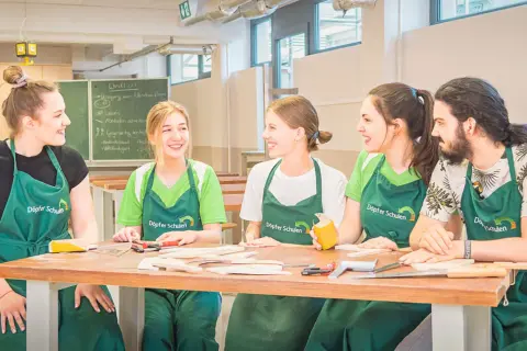 Fünf junge Menschen in grünen Schürzen lachen und arbeiten an einem Holztisch. Hier beginnt ihre kostenfreie Ausbildung Ergotherapie.