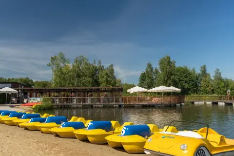 Żółte rowerki wodne czekają na plaży nad zalewem żyrardowskim. W tle drewniana kładka i zielone drzewa.