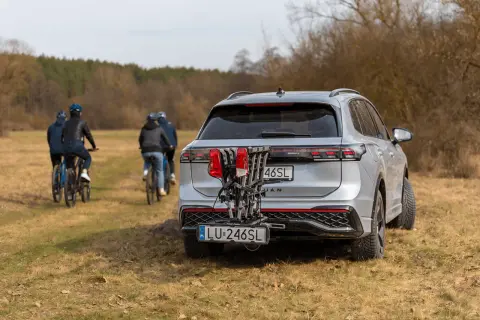 Srebrny SUV z rowerami na bagażniku i dodatkową tablicą rejestracyjną LU-246SL. W tle cztery osoby na rowerach.