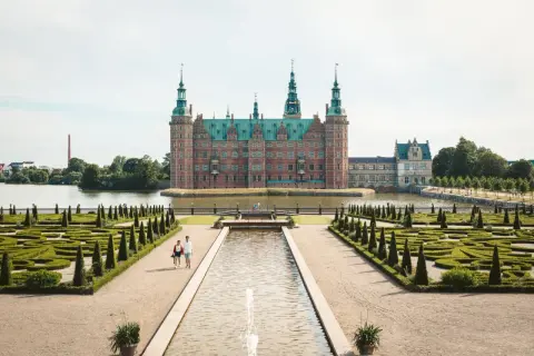 Ett imponerande slott i Danmark, omgivet av vatten och med en vacker barockträdgård. Ett par promenerar längs en damm framför slottet.