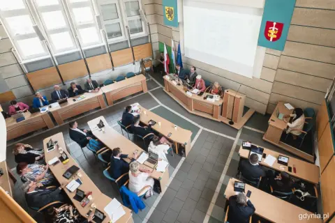 Obrady rady miasta, podczas których odbywa się udzielenie absolutorium. Ludzie siedzą przy stołach, niektórzy pracują na laptopach.