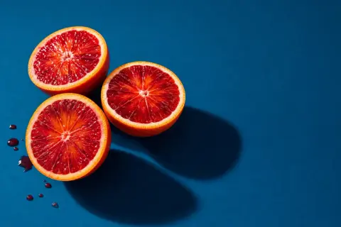 Vibrant fruit photography: three blood oranges halved, revealing their deep red flesh, with a few juice droplets on a deep blue background.