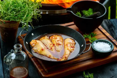 Kycklingvingar i en glansig, kryddig egen marinad i en stekpanna, redo för grillen. Bredvid finns färska örter och flingsalt.