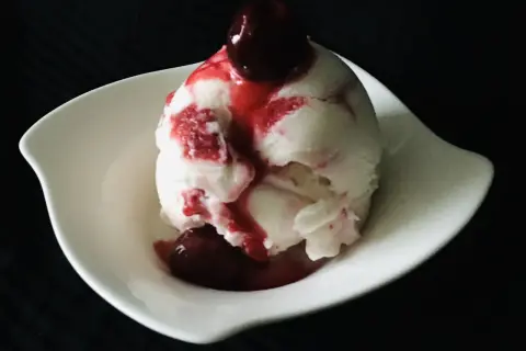 Ein Löffel cremiges Sauereis mit Kirschsauce und einer ganzen Kirsche obendrauf, serviert in einer weißen Schale.