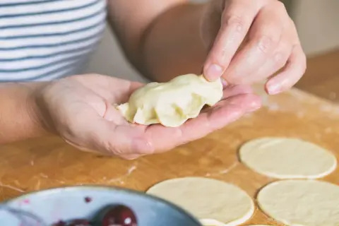 Dłonie formują pieroga z ciasta na pierogi bez jajka. Na blacie leżą okrągłe placki ciasta.