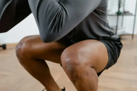 Ein Mann macht Kniebeugen, seine Knie sind gebeugt. Er hat leichte Narben auf dem rechten Knie, die auf frühere Schmerzen in der Kniekehle hindeuten könnten.