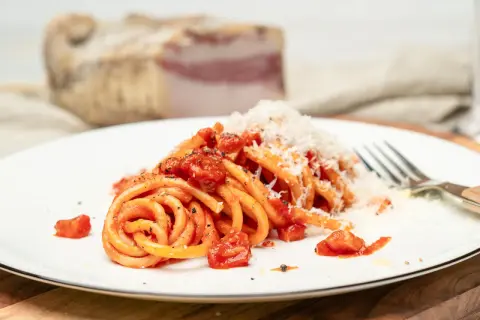 Ein Teller Pasta all'amatriciana, mit Tomaten und geriebenem Käse bestreut. Im Hintergrund ein Stück Guanciale, die Zutat für das authentische amatriciana rezept.