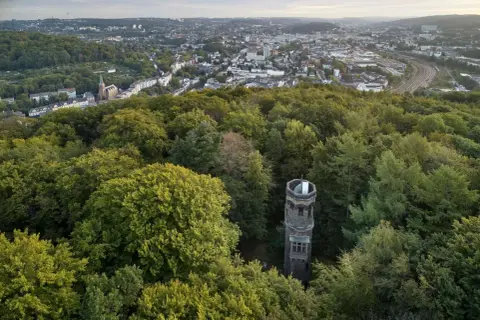 Der von der Heydt Turm ragt aus einem dichten grünen Wald hervor, mit einer Stadt im Hintergrund.