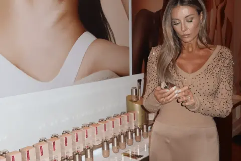 Kobieta z długimi blond włosami trzyma buteleczkę Clarins Double Serum Foundation. Na półce stoją dziesiątki opakowań tego podkładu.