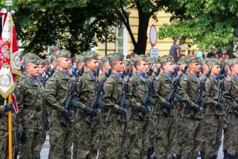 Żołnierze w mundurach defilują z karabinami. W tle sztandar i drzewa. Kategoria e wojsko.
