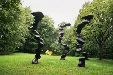 Drei schwarze, verdrehte Skulpturen stehen im **Skulpturenpark Wuppertal** auf einer grünen Wiese vor Bäumen. Eine gelbe Skulptur liegt im Hintergrund.