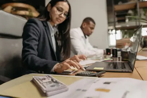 Kobieta w okularach liczy na kalkulatorze, obok stos banknotów i laptop.