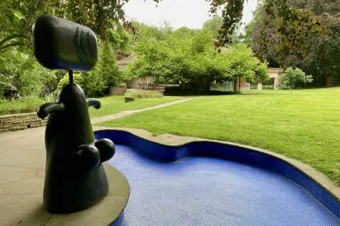 Eine abstrakte schwarze Skulptur steht neben einem blauen Mosaikbecken in einem **Skulpturenpark**. Im Hintergrund ein gepflegter Rasen und Bäume.