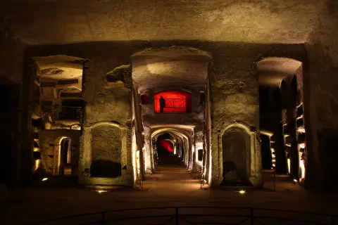 Corridoio sotterraneo illuminato, con nicchie scavate nella roccia e un'apertura rossa in lontananza, evocando l'atmosfera della tomba di San Gennaro.