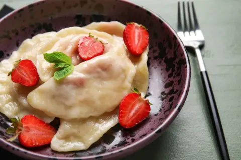 Pyszne pierogi z truskawkami, udekorowane świeżymi owocami i miętą, czekają w misce na widelcu.