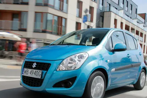 Błękitny Suzuki Splash jedzie ulicą miasta. W tle nowoczesne budynki i ludzie.