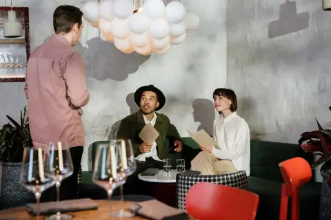Kelner w różowej koszuli rozmawia z parą przy stoliku. Zastanawiają się, ile zarabia kelner, przeglądając menu.
