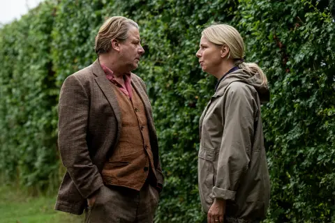 Två personer står och pratar intensivt framför en grön häck. Mannen bär kavaj och väst, kvinnan en jacka. En scen från en dansk kriminalserie på TV4.