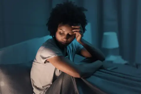 A woman sits in bed, head in hand, eyes closed, appearing to be experiencing fragmented sleep causes.