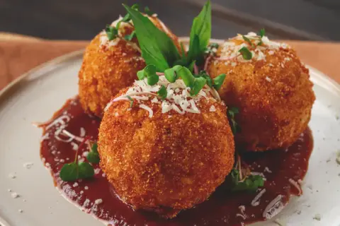 Trois arancini, spécialité italienne, dorés et croustillants, servis sur une sauce rouge avec des herbes fraîches et du parmesan râpé.