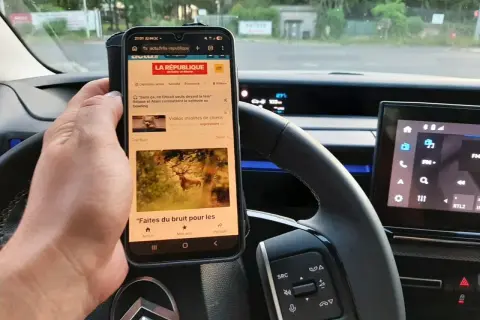 Main tenant un téléphone au volant, risque d'amende. L'écran affiche des articles d'actualité, un danger pour la conduite.