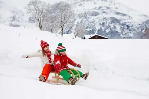Zwei Kinder rodeln fröhlich durch den Schnee. Ein idyllisches Bild vom Winter in der Schweiz mit verschneiten Bergen und einem kleinen Haus im Hintergrund.