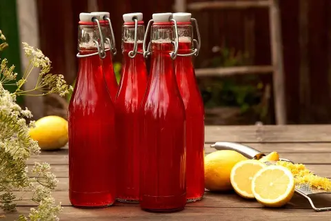 Fyra flaskor med klar, röd rabarbersaft står på ett träbord. Bredvid finns citroner och ett rivjärn. En riktig saftmaja-dröm!