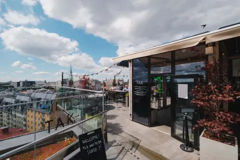 En takterrass med uteserveringar i Stockholm. Soliga moln på himlen, folk sitter vid borden och njuter av utsikten.