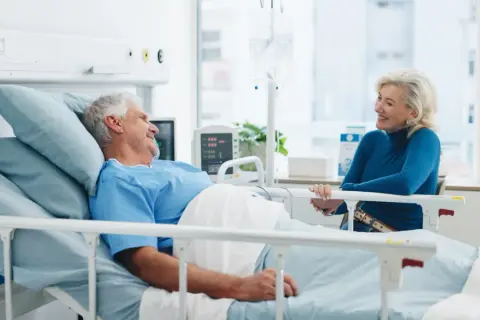 Un homme âgé, en chemise d'hôpital, sourit à une femme assise à son chevet. Une scène réconfortante d'apa et d'hospitalisation.
