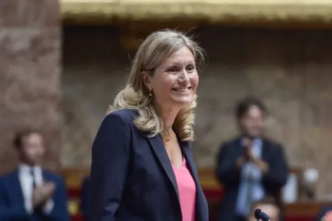 Une femme souriante, peut-être Vianney Pivet, d'origine inconnue, porte un blazer rayé et un haut rose.