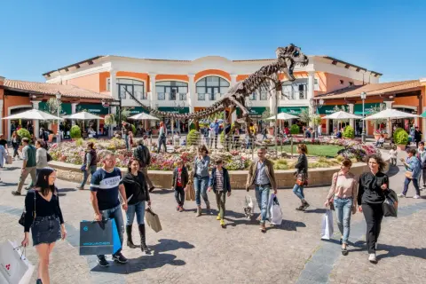 Un T-Rex scheletrico domina il centro del campo rom Castel Romano, circondato da negozi e gente che fa shopping.
