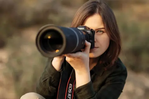 Kobieta z aparatem Canon EOS R7 uczy się, jak robić zdjęcia. Skupiona na kadrze, z obiektywem skierowanym w stronę widza.