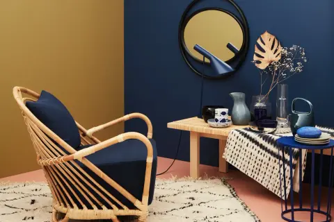 Ein Rattansessel mit dunkelblauem Kissen steht auf einem flauschigen Teppich. Daneben ein Tisch mit Deko. Welche Farbe passt zu Gold? Dunkelblau und Senfgelb harmonieren perfekt.