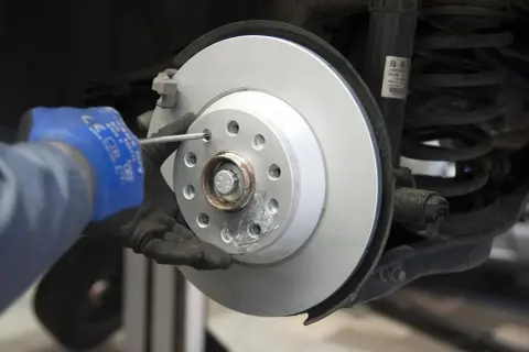 Mechanik w niebieskiej rękawicy dokręca śrubę podczas wymiany tarcz hamulcowych. Widać nowy, lśniący dysk hamulcowy i sprężynę amortyzatora.