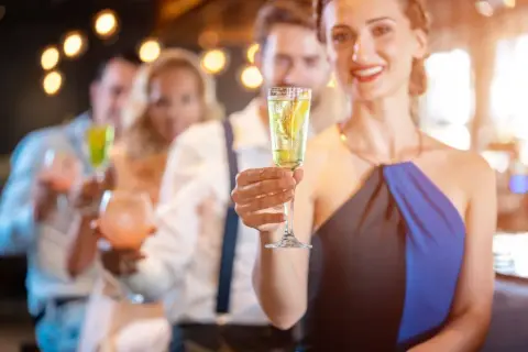 Uśmiechnięta kobieta trzyma kieliszek z drinkiem z prosecco i wódką, w tle bawią się inni goście.