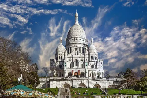 Bazylika Sacré-Cœur na wzgórzu Montmartre w Paryżu, z karuzelą w tle i dramatycznym niebem.