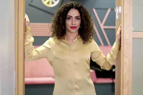 Helya Riazat, med lockigt mörkt hår och rött läppstift, står i en dörröppning i en guldig blus.