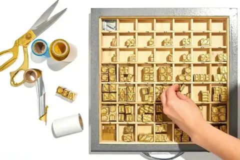 A hand selects brass letter stamps from a tray, ready to create a monogram. Gold scissors and foil rolls are nearby.