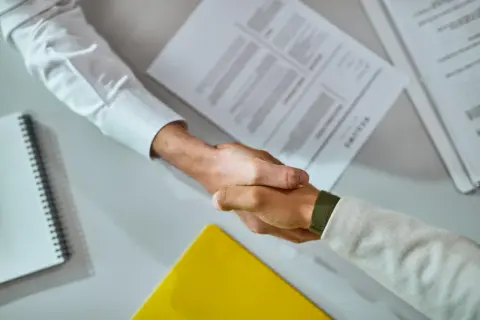 Uścisk dłoni po podpisaniu umowy stażowej. Na stole dokumenty i notatnik.