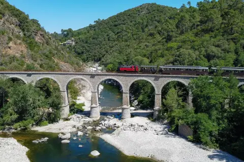 Le petit train des Cévennes traverse un viaduc de pierre au-dessus d'une rivière. La durée du trajet est une invitation à la découverte de ce paysage verdoyant.