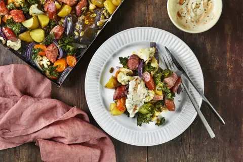 En tallrik med ugnsrostade grönsaker och korv, ett perfekt potatis recept. Bredvid syns en ugnsplåt med samma läckra innehåll.