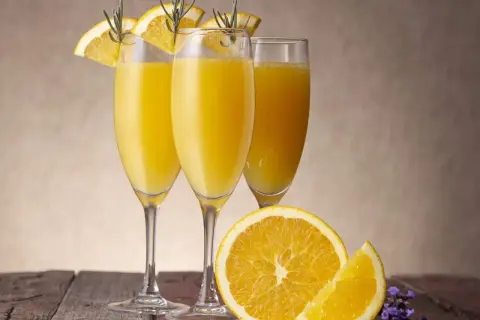 Tre glas med en gyllene drink med cava, garnerade med apelsinskivor och rosmarin. En apelsin och lavendel på träbordet.