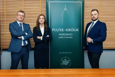Trzy osoby w garniturach stoją przed zielonym banerem kancelarii Piatek-Krolik.