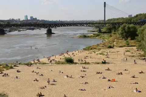 Słoneczna poniatówka nad Wisłą. Ludzie opalają się na piaszczystej plaży, w tle most i panorama Warszawy.