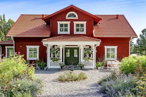 Vackert rött hus med vita detaljer, en veranda och en blomstrande trädgård. Ett perfekt exempel på att **bygga hus i gammal stil**.