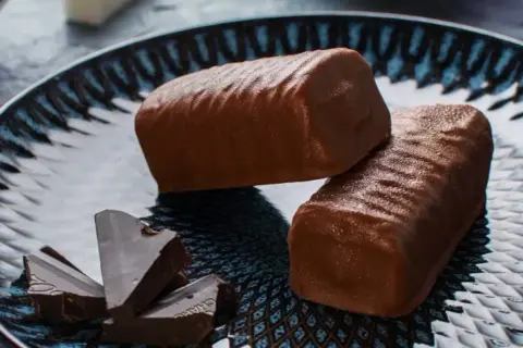 Dwa czekoladowe batony proteinowe na talerzu, obok kawałki gorzkiej czekolady. Idealne do domowego batony proteinowe przepis.