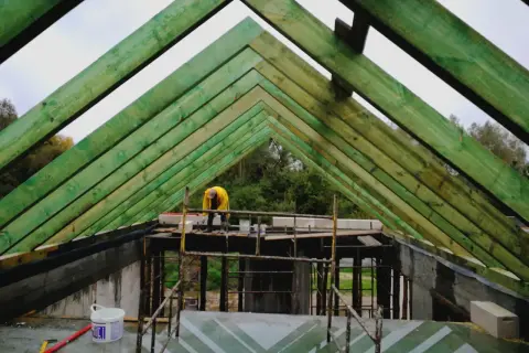Zielone elementy więźby dachowej tworzą konstrukcję dachu, nad którą pracuje budowlaniec w żółtej kurtce.